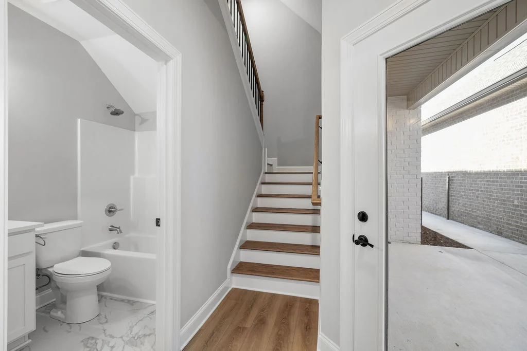 New Home Chattanooga TN. Bright entryway by Pratt Home Builders features a wooden staircase, a bathroom with white fixtures, and access to a covered porch.