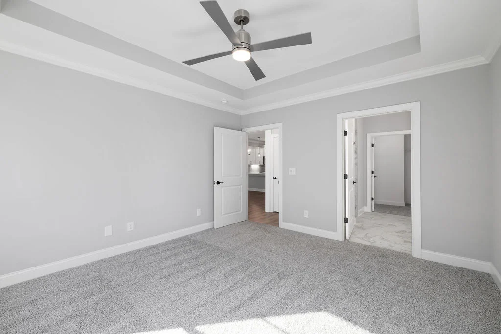 New Home Chattanooga TN. Spacious gray bedroom with tray ceiling, fan, carpet, and two doors leading to a hallway and bathroom. Built by Pratt Home Builders.