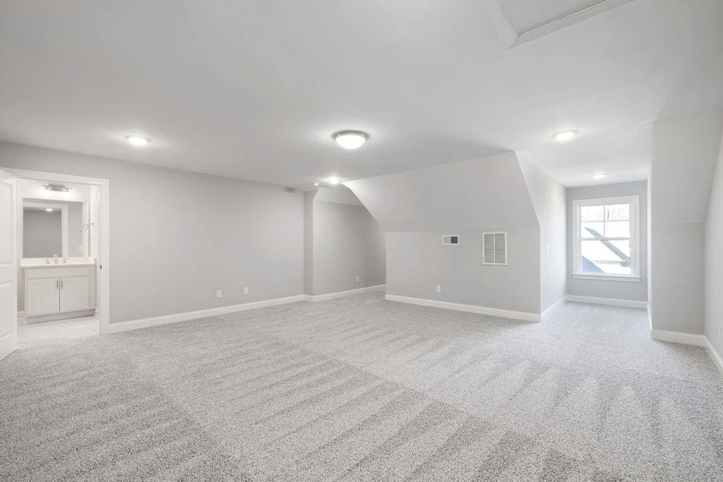 Spacious, carpeted attic room with sloped ceiling, small window, and adjacent bathroom in a Pratt Home Builders property in Chattanooga, TN.