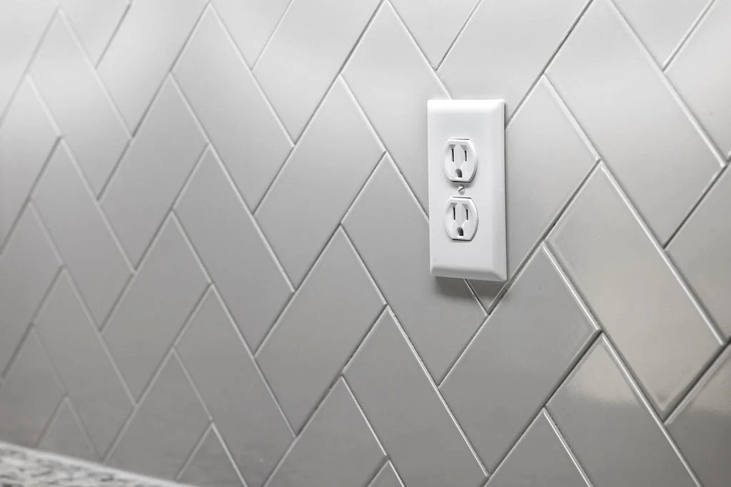White herringbone tile backsplash with a white outlet cover. Design by Pratt Home Builders in Chattanooga TN.