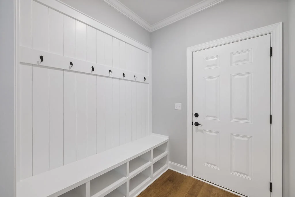 Entryway with white bench, storage cubbies, and hooks on paneled wall by Pratt Home Builders in Chattanooga TN. White door with black handle.