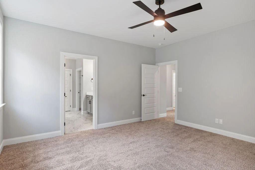 Spacious, carpeted bedroom with gray walls, ceiling fan, and two open doors leading to a bathroom. Pratt Home Builders in Chattanooga TN.