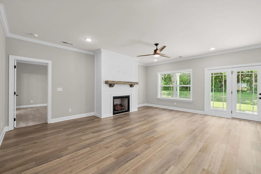 Spacious living room by Pratt Home Builders in Chattanooga, TN, featuring hardwood floors, white fireplace, ceiling fan, and garden view through large