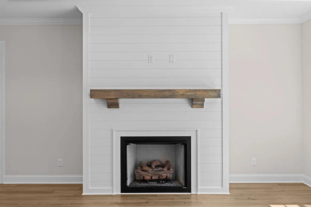 White shiplap fireplace with wooden mantel and brick hearth by Pratt Home Builders in Chattanooga TN, set against beige walls and wooden floors.